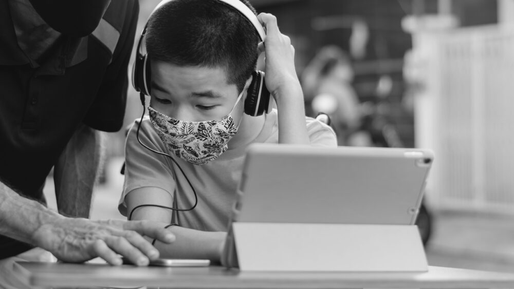 Black and white photo of student receiving help from a teacher. He's working on a laptop while wearing headphones and mask..