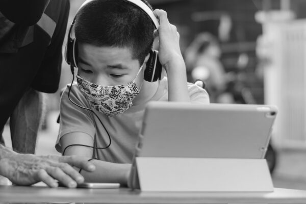 Black and white photo of student receiving help from a teacher. He's working on a laptop while wearing headphones and mask..