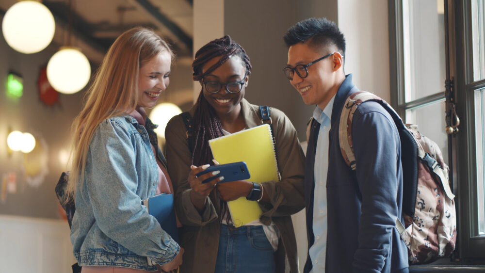 Happy young university students friends relaxing with smartphone in corridor. Multiethnic hipster college people watching video on smartphone and laughing resting after lecture in hallway