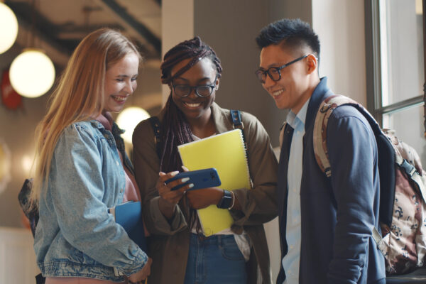 Happy young university students friends relaxing with smartphone in corridor. Multiethnic hipster college people watching video on smartphone and laughing resting after lecture in hallway