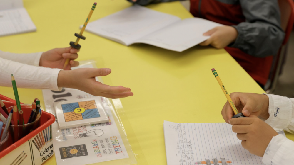 Table with student hands holding pencils and notebooks.