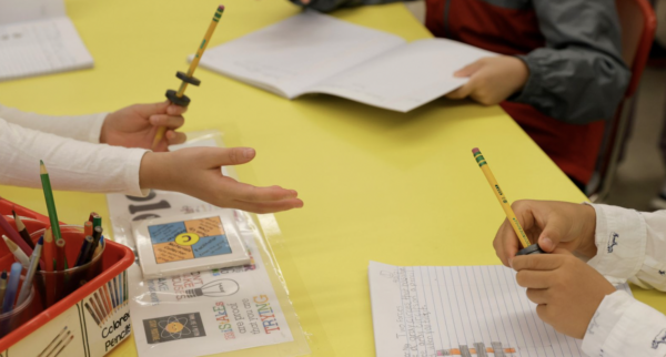 Table with student hands holding pencils and notebooks.