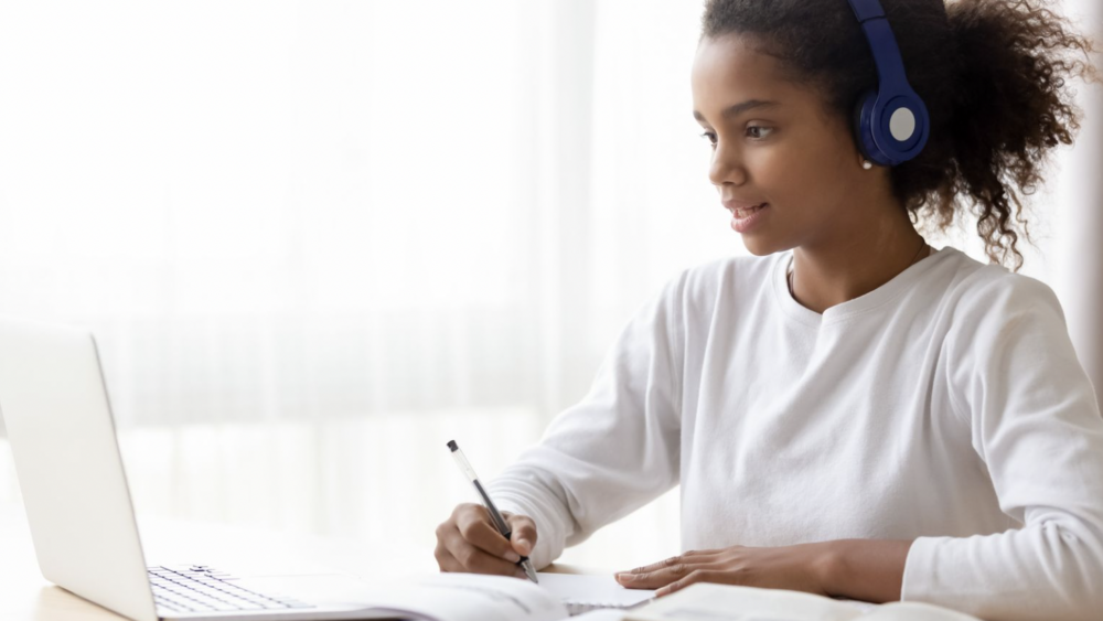 African-American girl wearing a white shirt and blue headphones. Taking notes while sitting at a laptop.