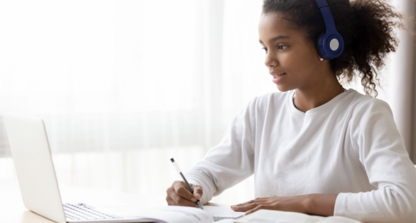 African-American girl wearing a white shirt and blue headphones. Taking notes while sitting at a laptop.