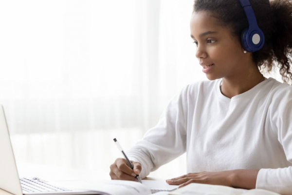 African-American girl wearing a white shirt and blue headphones. Taking notes while sitting at a laptop.