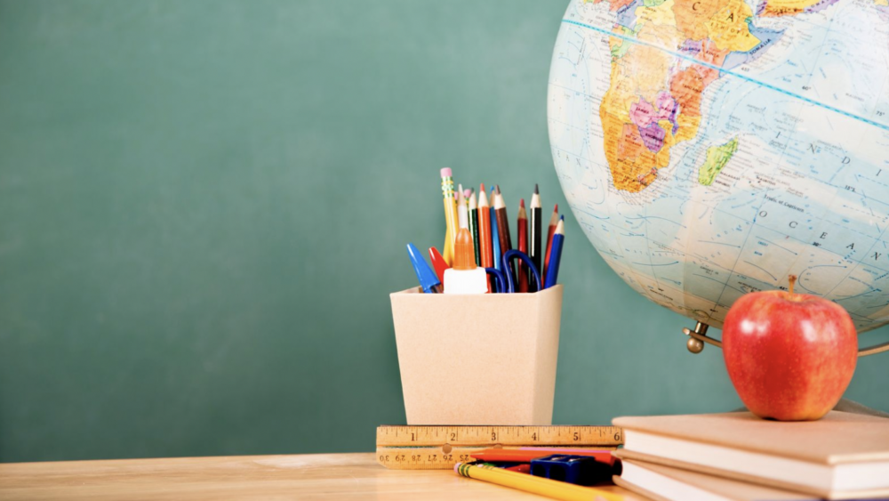 Green chalkboard with a globe, cup of pencils, books, and apple on a desk.