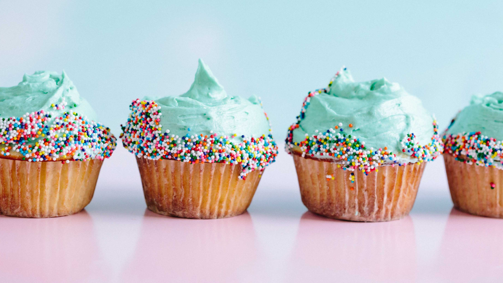 A line of cupcakes with aquamarine frosting and sprinkles against a pink and blue background