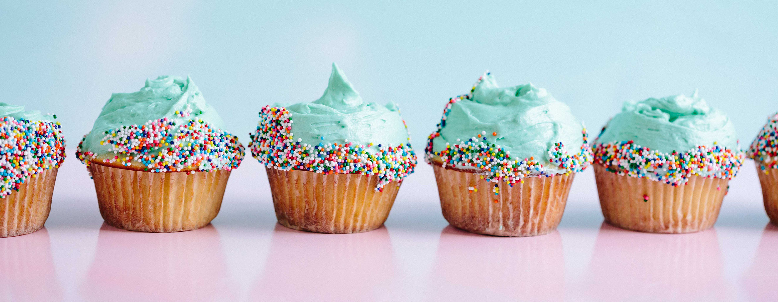 A line of cupcakes with aquamarine frosting and sprinkles against a pink and blue background