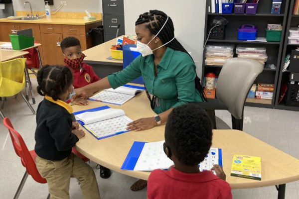 Teacher with a mask on working at a table with young students.