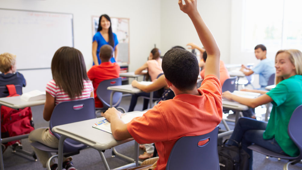 Student raising hand while sitting at a desk in a school classroom